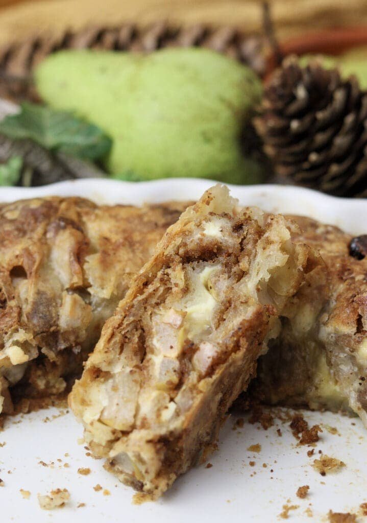 A slice of cheese strudel with pears, raisins and cinnamon, with fresh green pear in the background. 