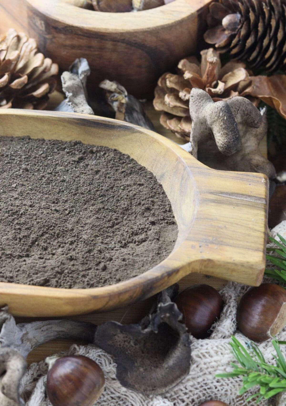 Dried black trumpet mushroom, turned into black trumpet powder in a wooden bowl with with black trumpet mushrooms, chestnuts and pine cones in the background.