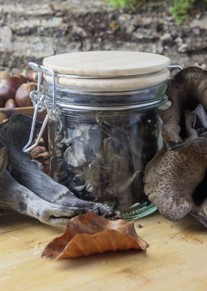 Dried black trumpets in a glass jar with fresh mushrooms and autumn leaves in the background