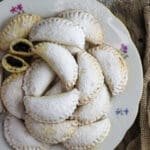 A plate of mouthwatering empanada cookies with one cookie cut in half where plum jam is visible and with a strain full of powdered sugar in the background