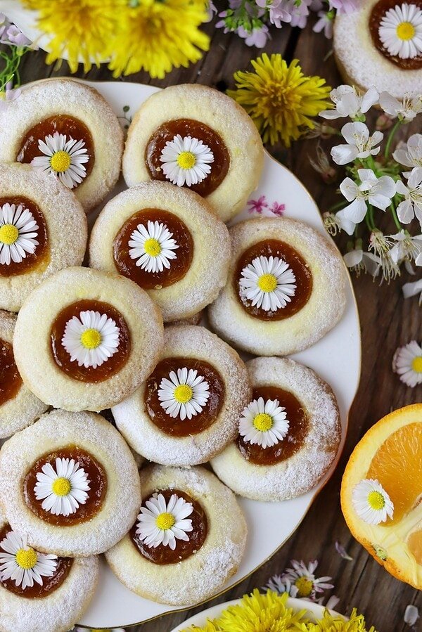 Golden dandelion thumbprint cookies with apricot jam on a white ceramic plate surrounded by spring flowers.