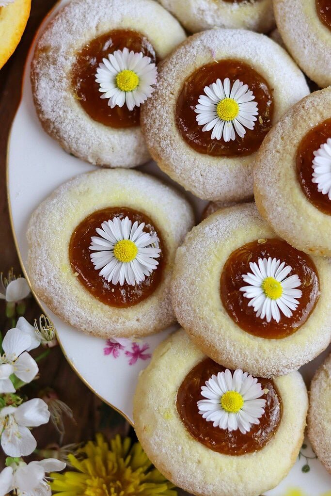 A close shot of dandelion cookies with apricot jam on a white plate with flowers in the background.