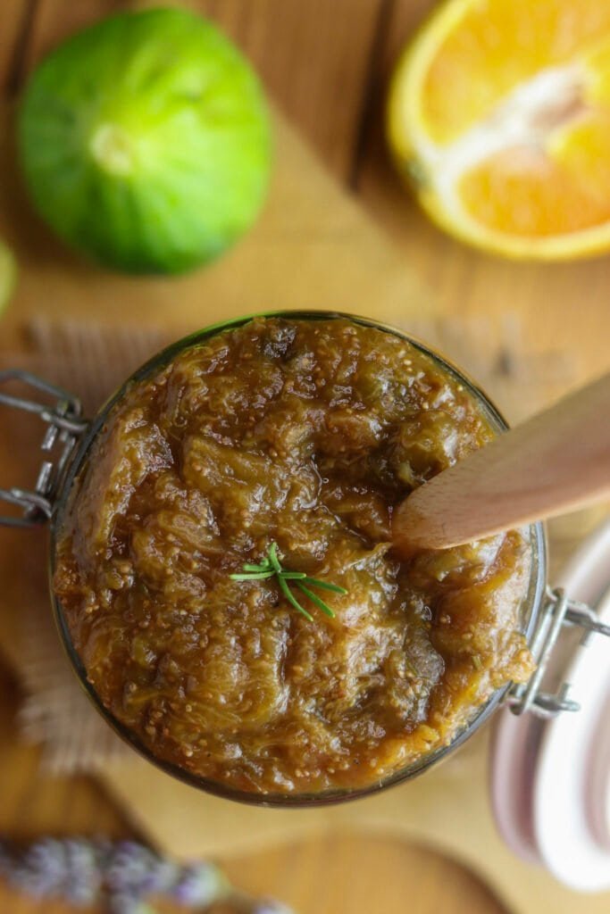Low sugar fig jam recipe, jar of jam with rosemary, with a fresh fig and orange half in the background
