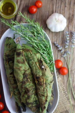 Grilled mackerels, wrapped in fig leaves and grilled with a rosemary sprig and tomato, olive oil and garlic in the background.