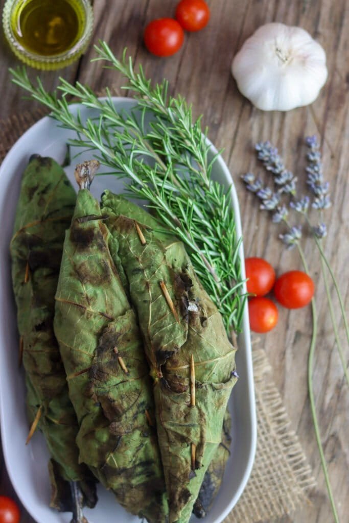 Grilled fig leeaf wrapped chub mackerels on a white plate with a rosemary sprig and garlic, tomato, lavender and olive oil in the background.