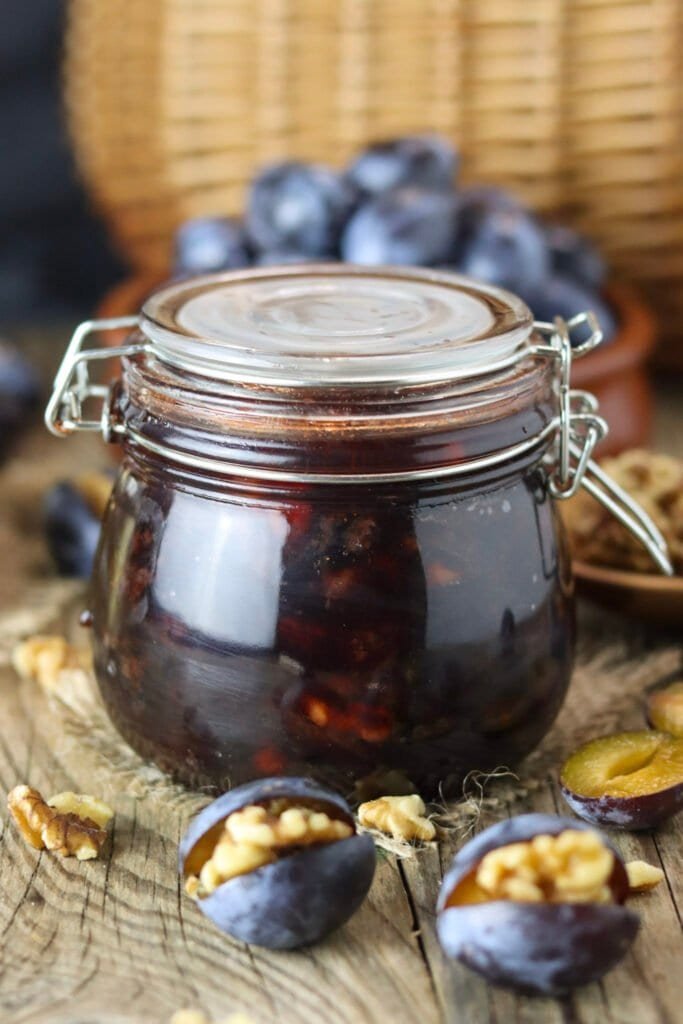 A closed jar of honey preserve with roasted plums and walnuts, with stuffed plums in the background.