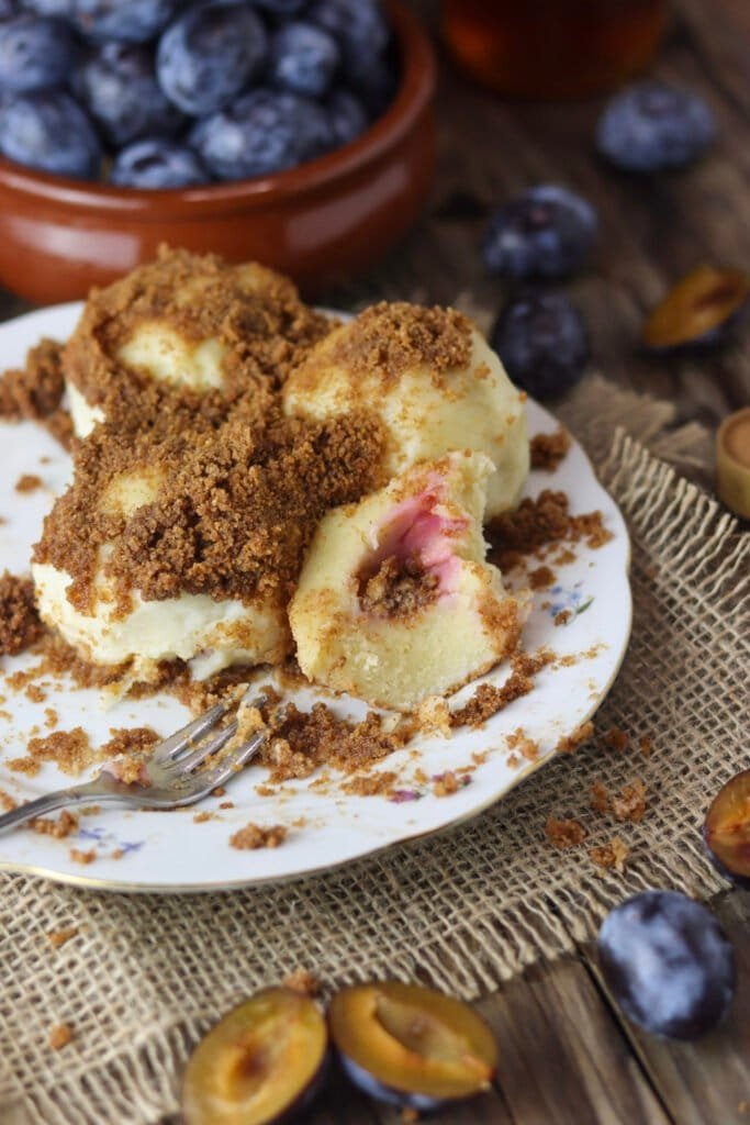 A plate loaded with plum dumplings, coated with breadcrumbs, a few already eaten, a bowl of plums in the background.