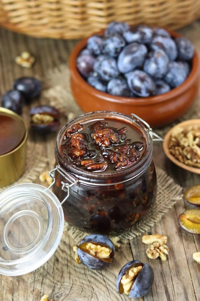 A jar of honey preserves with plums and walnuts, with fresh plums, stuffed plums and walnuts in the background.