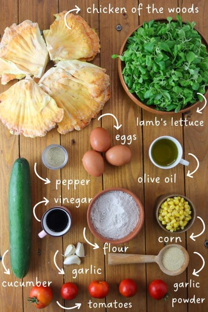Ingredients for a delicious fried chicken of the woods mushroom salad on a wooden kitchen counter: fresh chicken of the woods mushrooms, fresh buckwheat flour, garlic, garlic powder, white pepper, cooked corn, cucumber, tomato, lamb's lettuce, eggs, olive oil and vinegar.