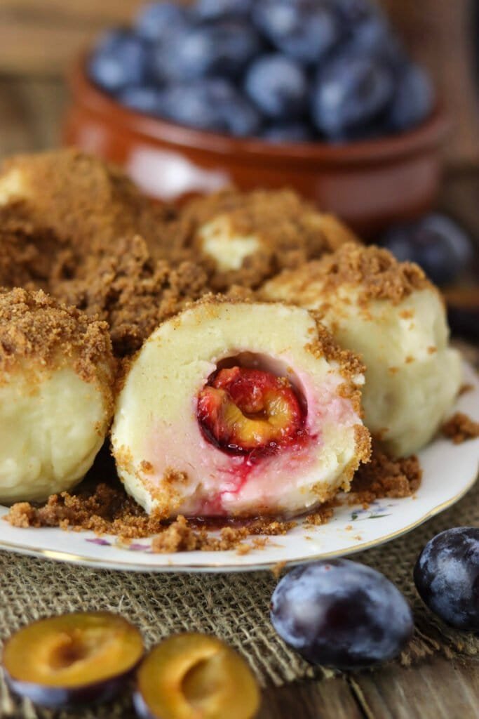 Dessert dumplings on a plate, made with cottage cheese, fresh plums, spelt flour and breacrumbs, with fresh plums in the background.