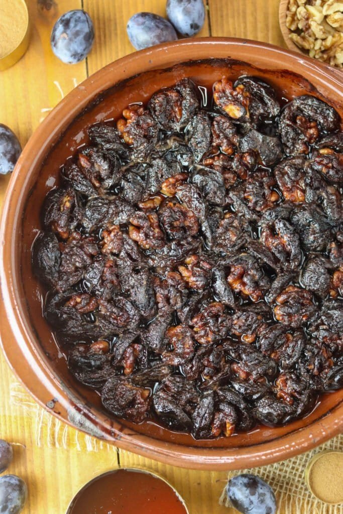 Baked plums with walnuts in a round baking pan, with honey, walnuts and fresh plums in the background.