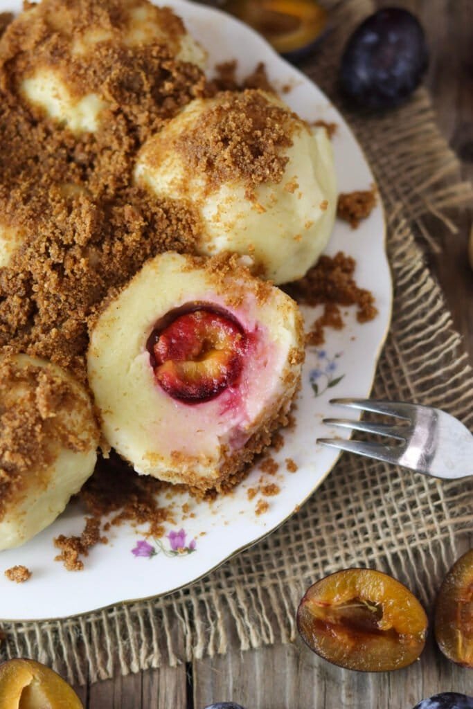 Spelt plum dumplings with cottage cheese on a rustic plate, covered with toasted breadcrumbs. Halved fresh plums in the background.