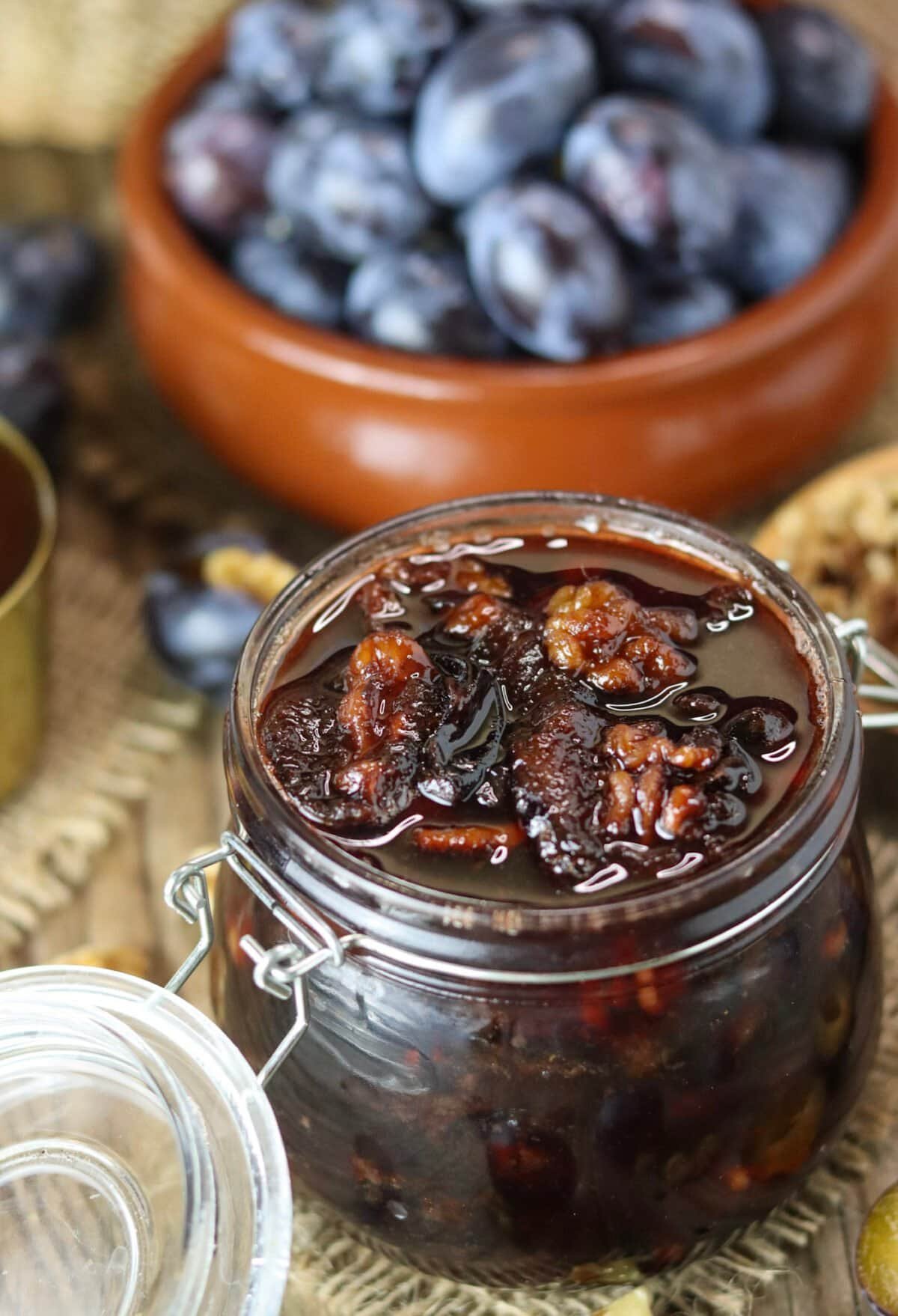 An open jar of walnut honey preserve with roasted plums on a rustic wooden table.