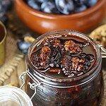 An open jar of walnut honey preserve with roasted plums on a rustic wooden table.