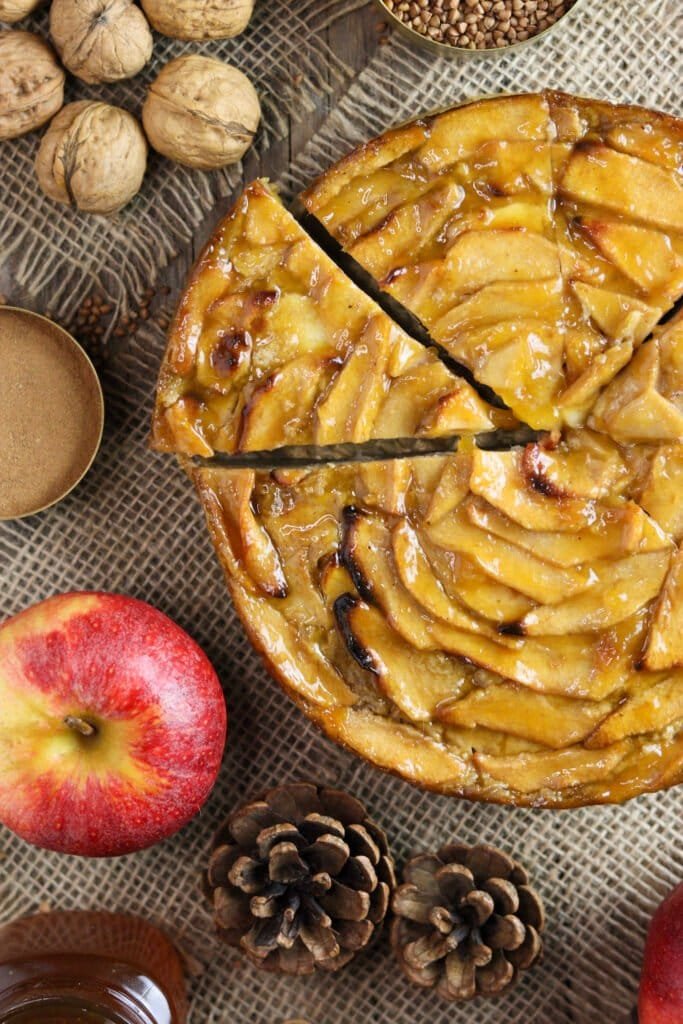 Sliced buckwheat flour cake on a rustic wooden table with a red apple, walnuts and two pine cones in the background.