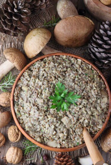 A top view of a rustic handmade bowl of buckwheat with fresh porcini mushrooms and a wooden spoon. Fresh porcini, walnuts and pine cones in the background.