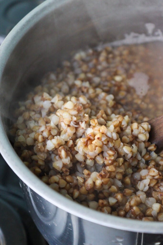 Buckwheat groats being cooked in a pot, one part lifted with a wooden spoon to show they are cooked enough.