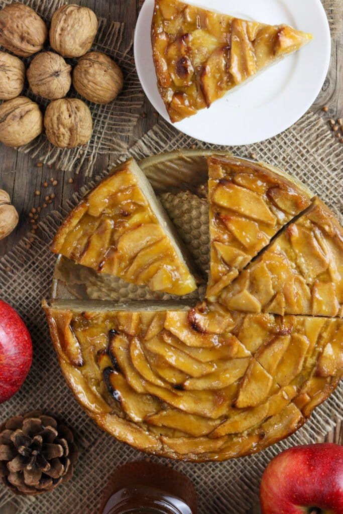 Sliced buckwheat apple cake on a baking sheet, one slice on a dessert plate with walnuts, apples and honey in the background.