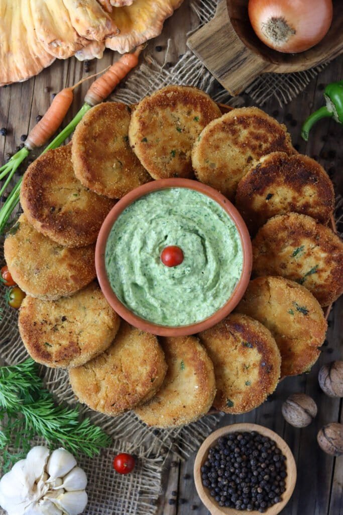 Chicken of the woods mushroom fritters with a green dip on a rustic plate, with an onion, garlic, black pepper, carrots, and walnuts in the background.