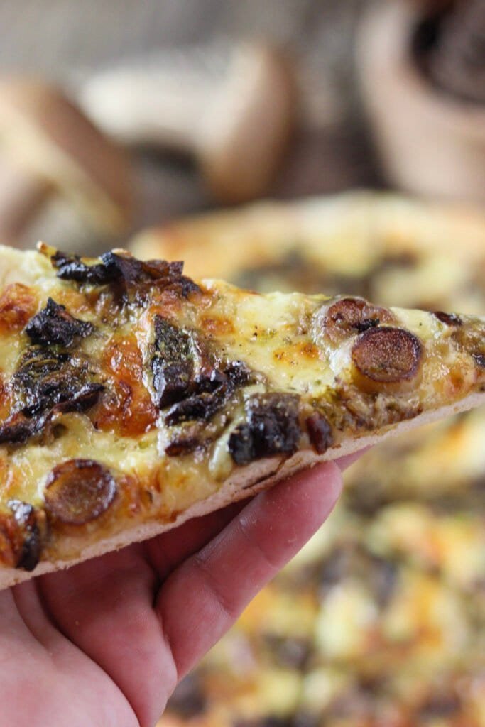 A slice of pizza with funghi porcini in my hand, with a whole pizza and fresh porcini in the background.