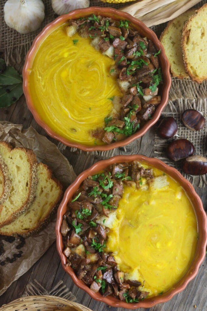 Two bowls saffron milk cap squash soup on a rustic wooden table with corn bread, garlic and chestnuts.