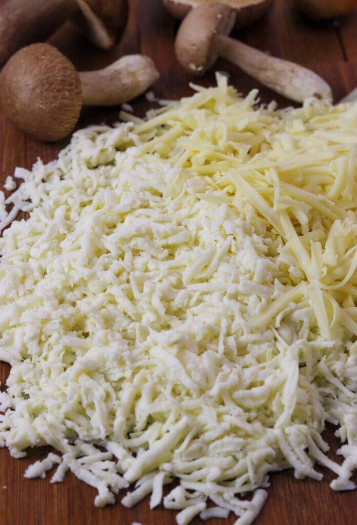 Shredded mozzarella cheese and cheddar on a wooden cutting board with porcini in the background.
