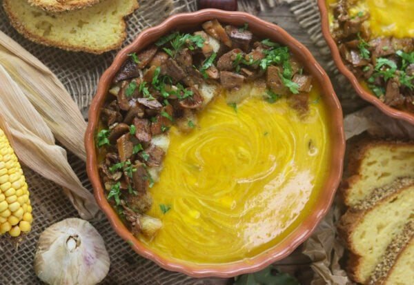 A rustic bowl with mushroom squash soup with saffron milk cap mushrooms, red kuri squash and bread croutons, with corn bread, dry corn and garlic head in the background.