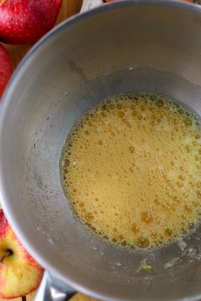 Eggs whisked in a large bowl with apples in the background.