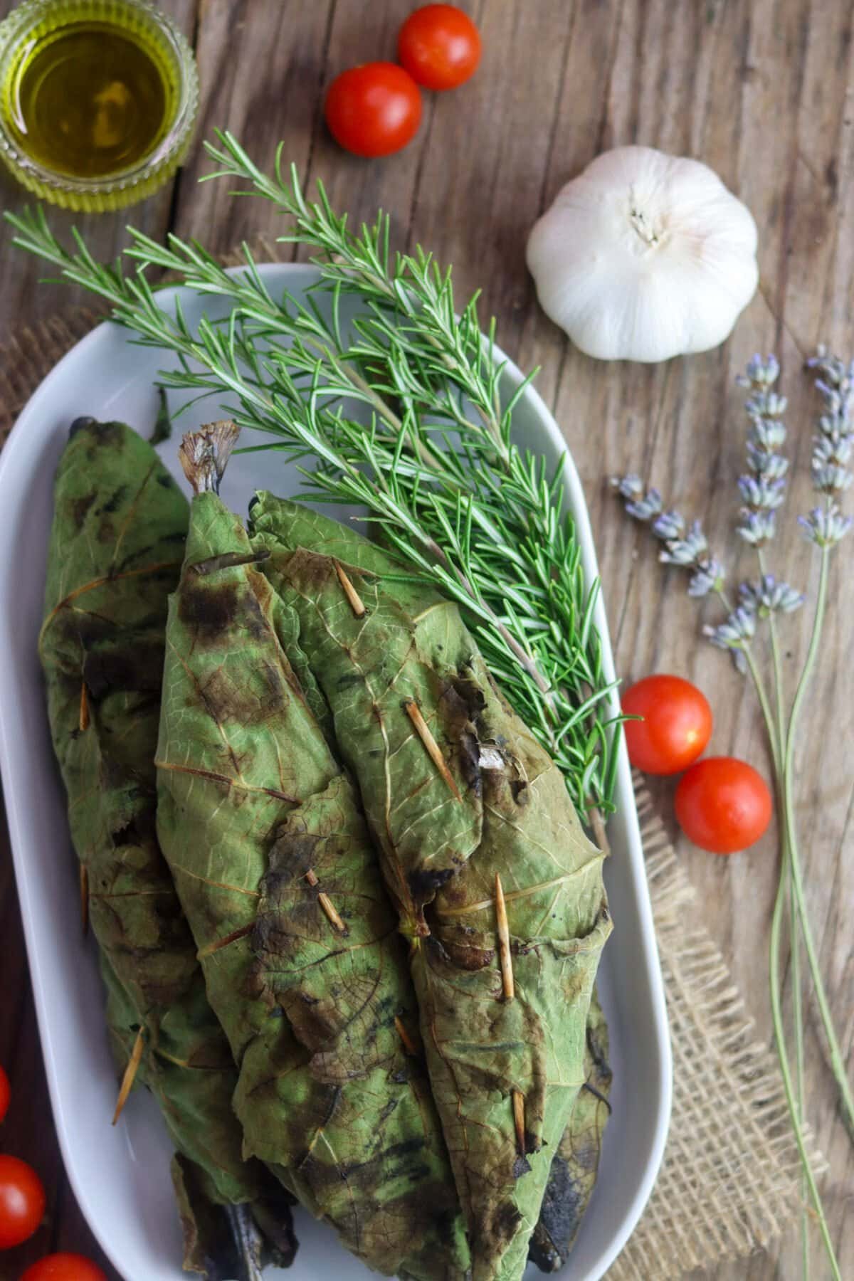 Grilled mackerels, wrapped in fig leaves and grilled with a rosemary sprig and tomato, olive oil and garlic in the background.