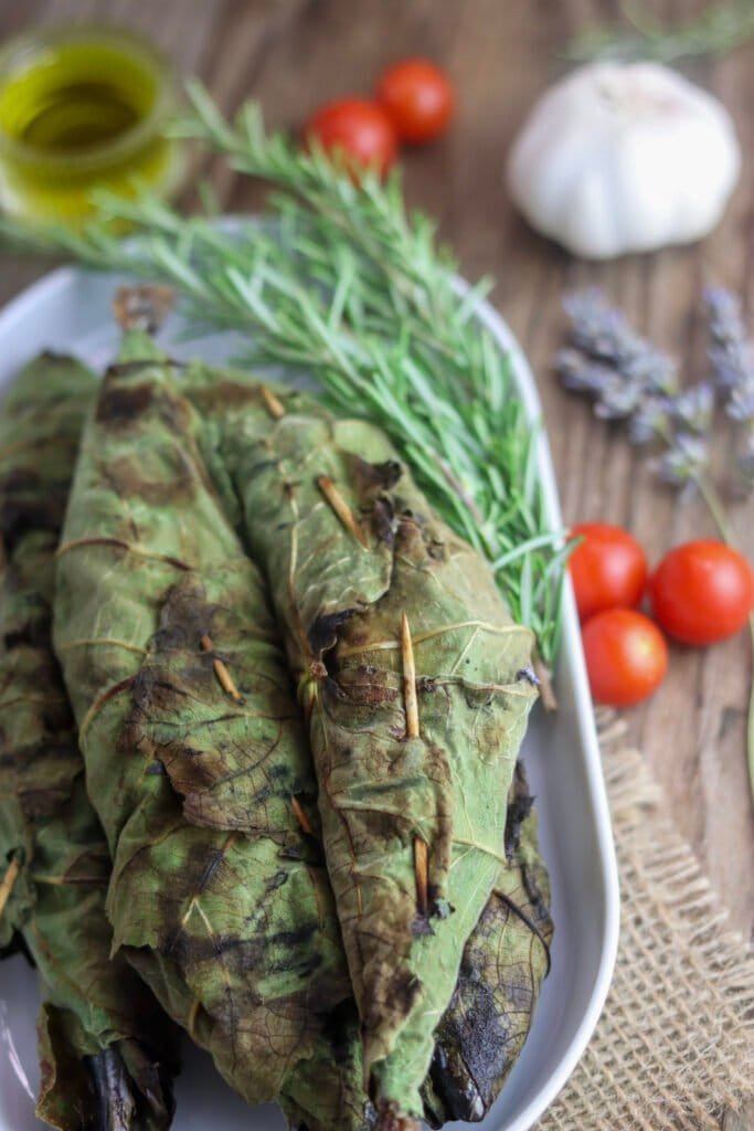 A stack of grilled chub mackerels, wrapped in fig leaves with rosemary, grilled, with tomato, olive oil and garlic in the background.