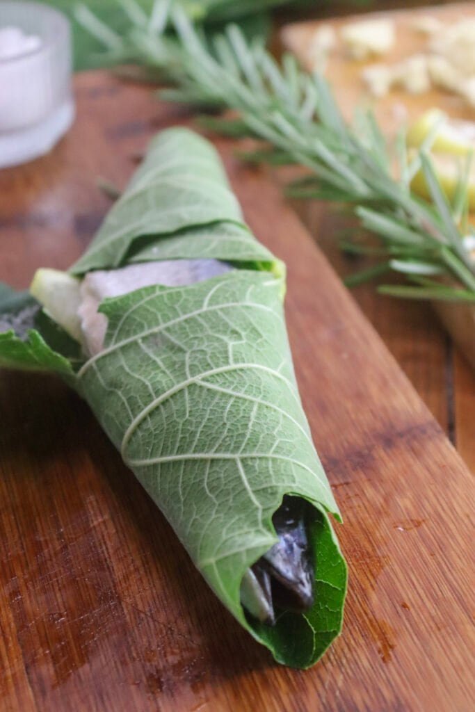Chub mackerel on a wooden plate, stuffed and wrapped in a large fig leaf, with rosemary in the background.