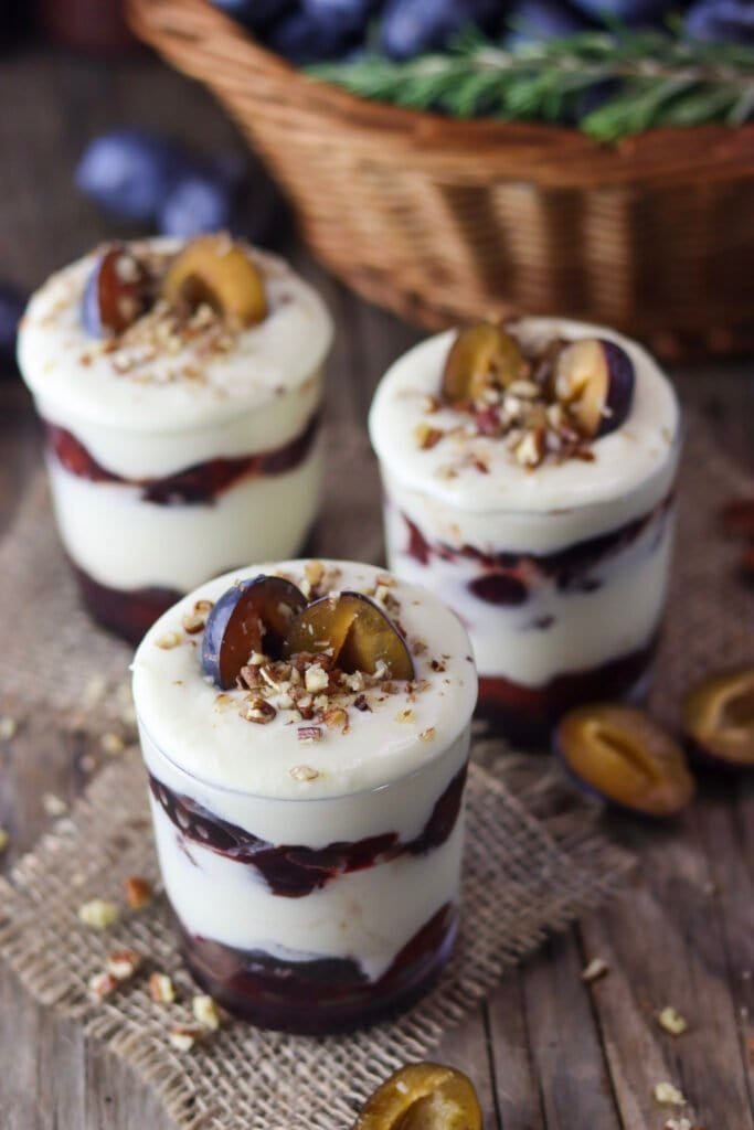 Three jars of delicious plum white chocolate parfait with a basket of plums, rosemary and ground pecans in the background.