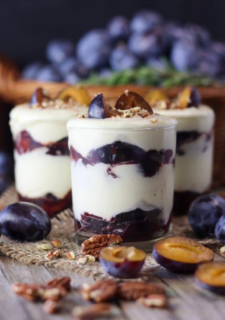 Three beautiful plum parfait jars on a wooden table, decorated with plums, with fresh plums and pecans around them and a full basket of plums in the back.